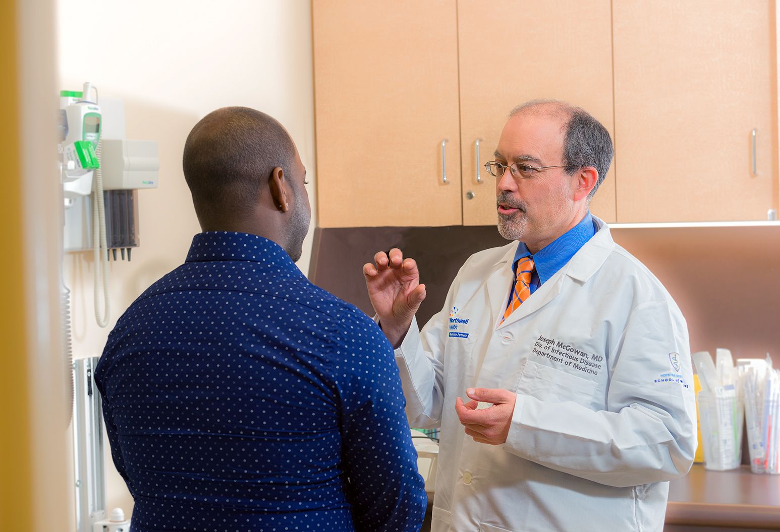 A male doctor in a white lab coat and glasses spends one-on-one time with a male patient, explaining something to him using hand gestures.